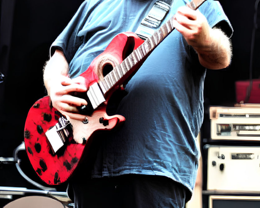White-haired musician plays red electric guitar on stage with sunglasses and beard.