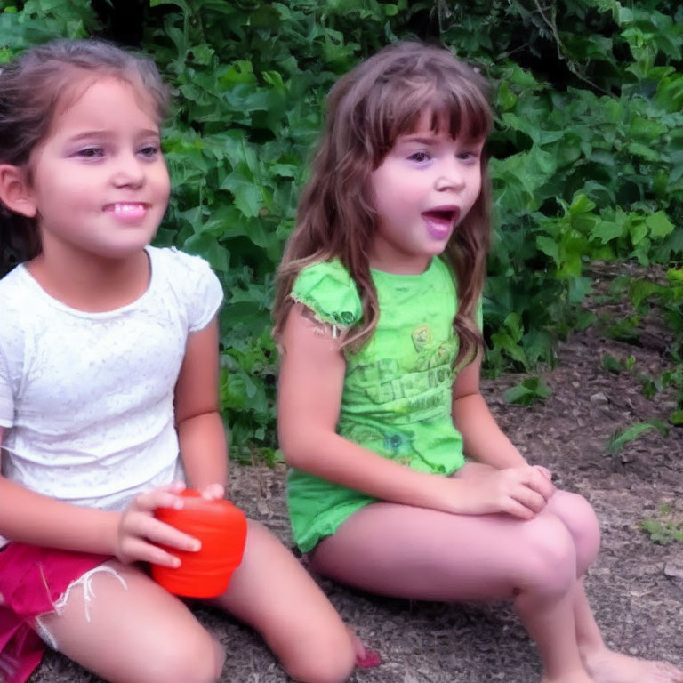 Two young girls sitting outside, one in green shirt with excited expression, the other smiling slightly.