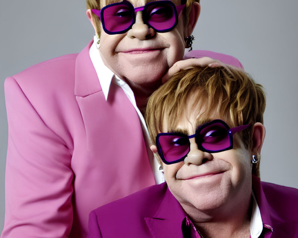 Two people in pink outfits and purple glasses pose against grey backdrop