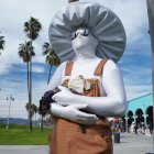White-faced figure statue with umbrella and apron against cloudy sky and palm trees