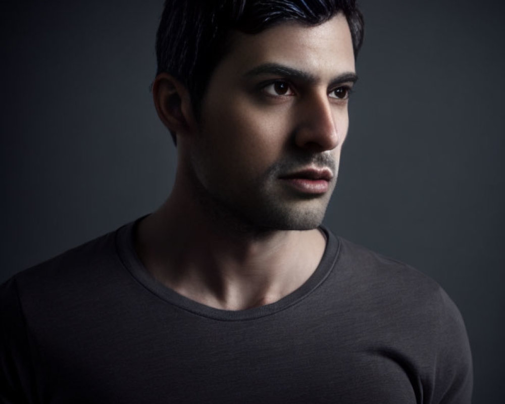Dark-haired man in black t-shirt with serious expression, looking sideways on dark backdrop.