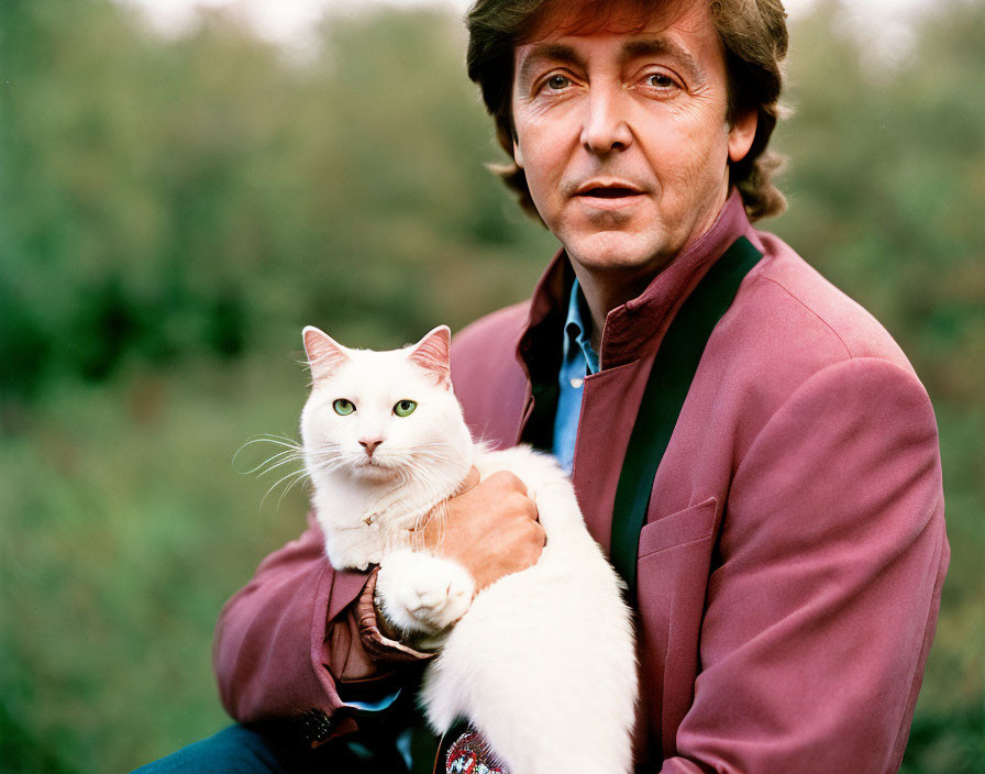 Brown-haired man in burgundy jacket holds white cat with green eyes in natural setting