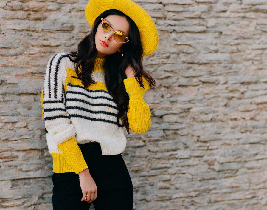 Fashionable individual in yellow hat and striped sweater against stone wall.