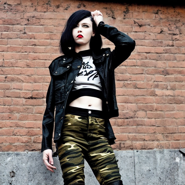 Black-haired woman in leather jacket against brick wall in cropped top and camo pants