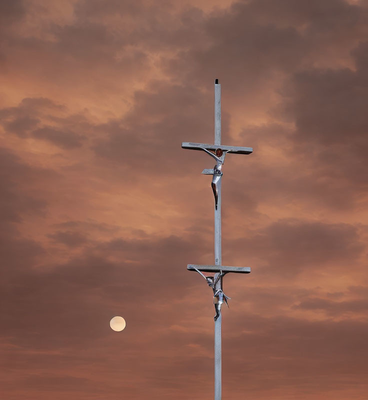 Metal figures on cross under orange dusk sky with white moon