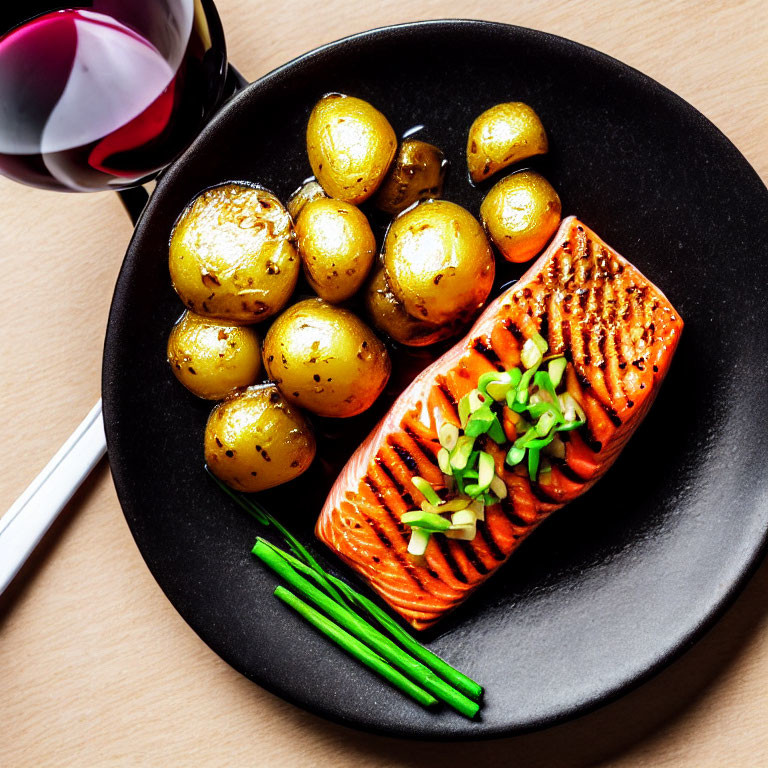Grilled salmon fillet and roasted potatoes with green onions on black plate with red wine.