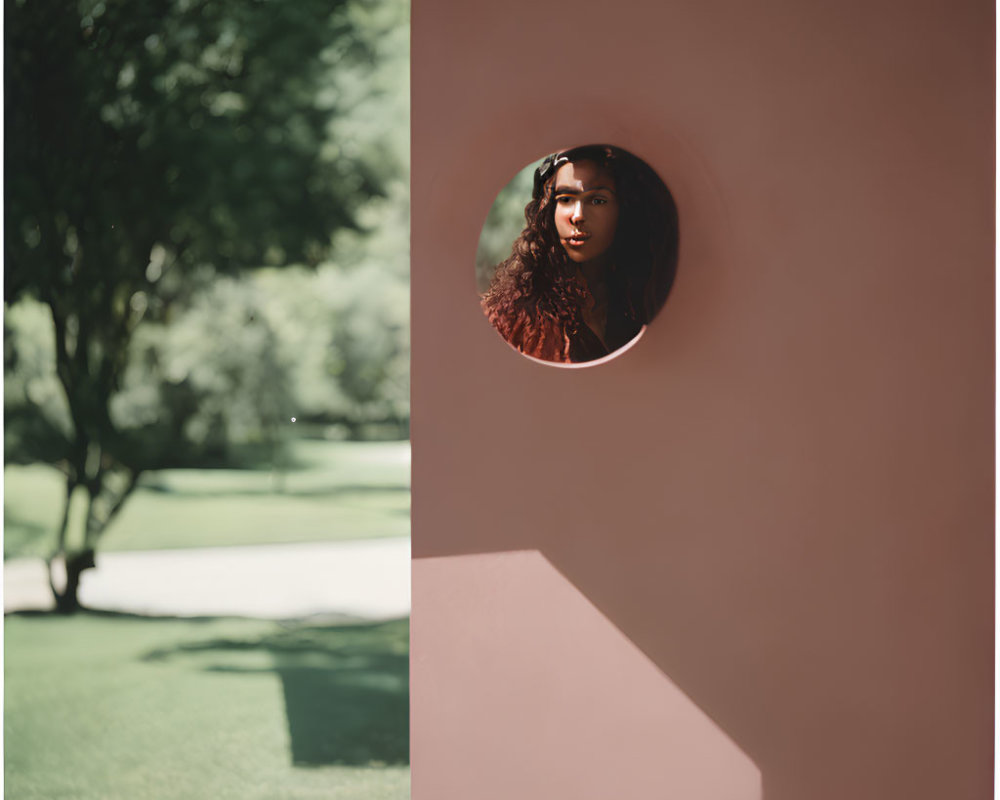 Woman looking through circular hole in pink wall with blurred green garden.