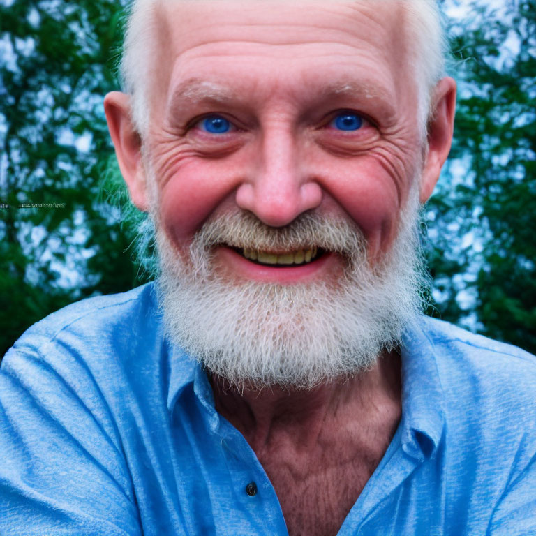 Elderly man with white beard and blue eyes smiling in blue shirt with tree background