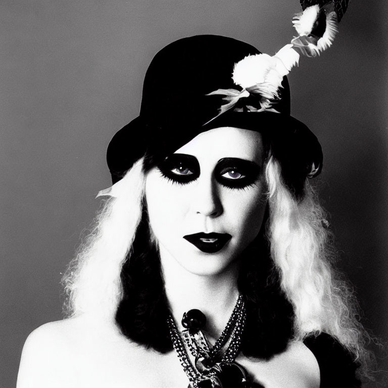 Monochrome image of person in dramatic makeup, feathered hat, layered necklaces, and curly hair