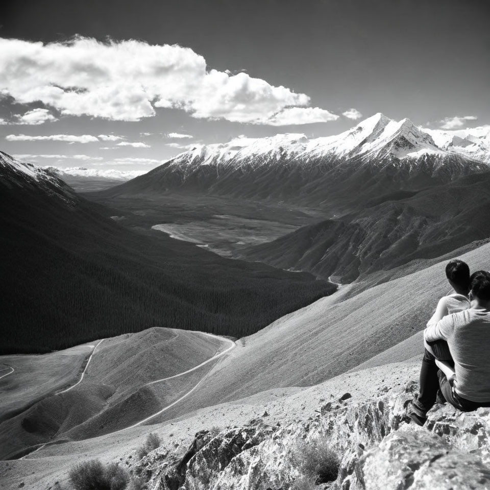 Two individuals on mountain edge with forested valleys and snowy peaks, black and white.
