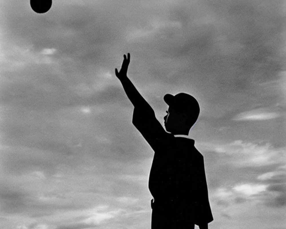 Child silhouette with baseball cap and glove reaching for ball in cloudy sky