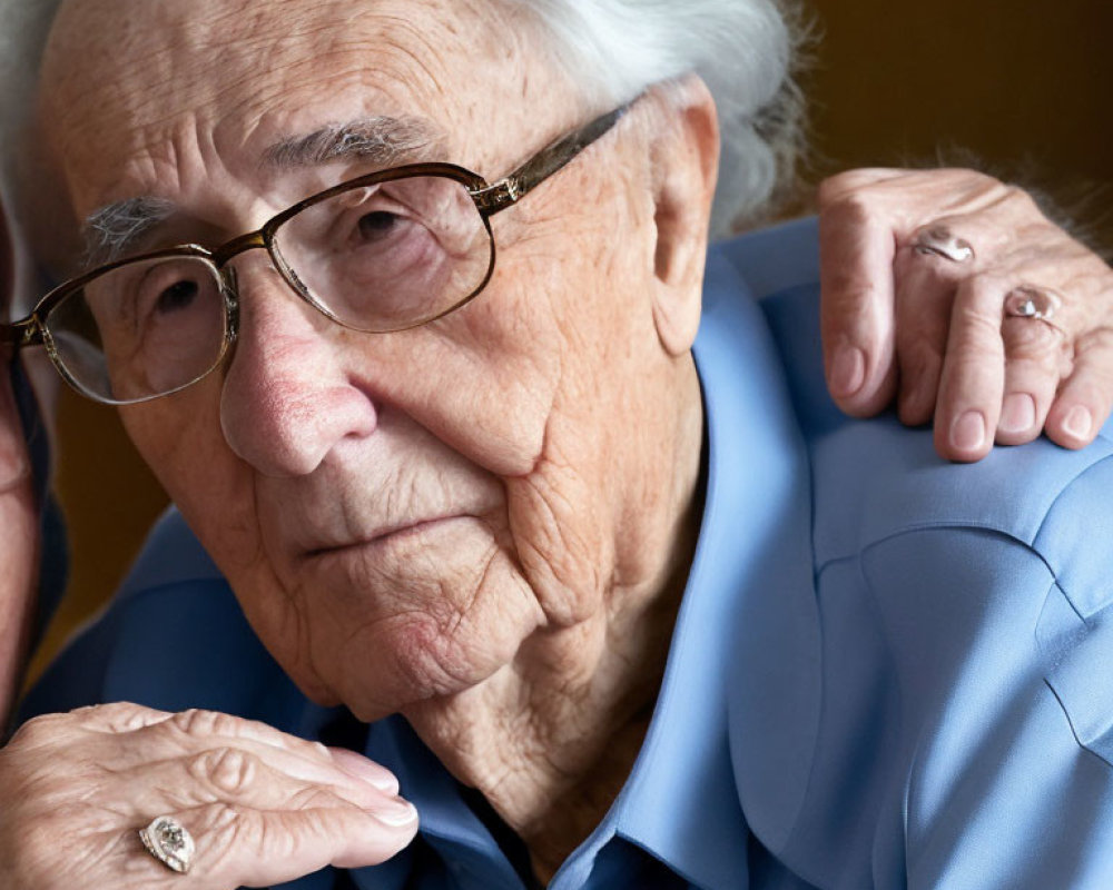 Elderly person in blue shirt with glasses, looking pensive with hand on shoulder