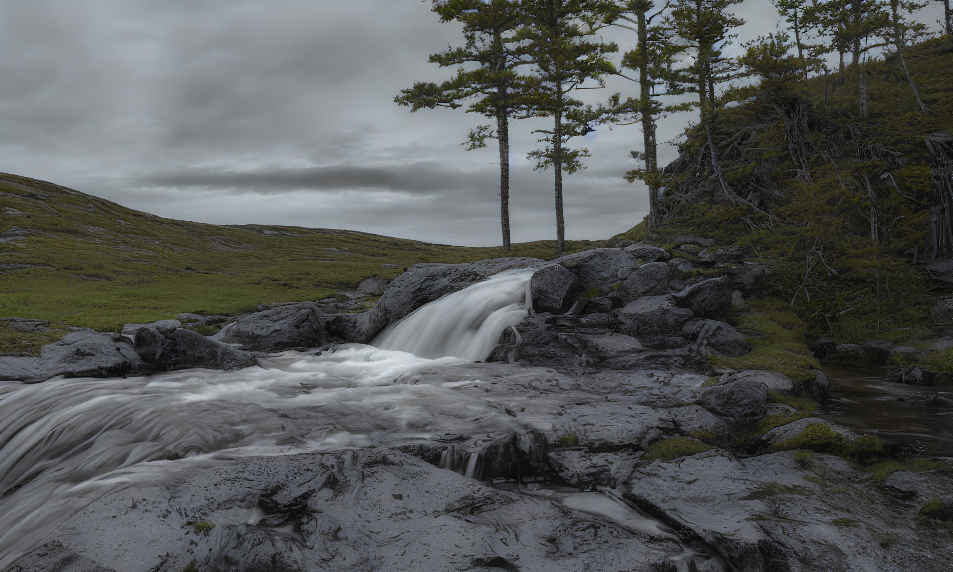 Tranquil waterfall scene with dark rocks, green grass, and cloudy sky