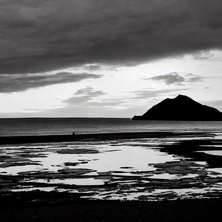 Monochromatic seascape with tidal pools, solitary figure, and hill under cloudy sky
