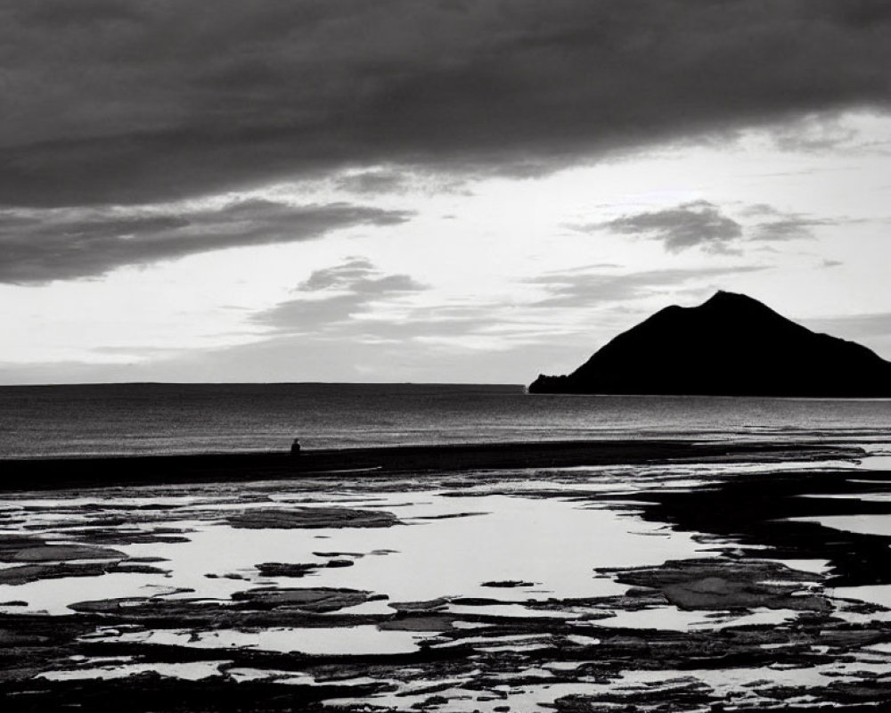 Monochromatic seascape with tidal pools, solitary figure, and hill under cloudy sky