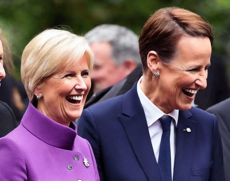 Two women in formal attire laughing: one in purple, the other in blue suit