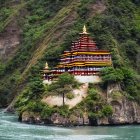 Golden-roofed multi-tiered temple on rocky hill near flowing river surrounded by lush green mountains