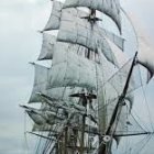 Tall ship with white sails, gold trim, and dark hull on cloudy sky.