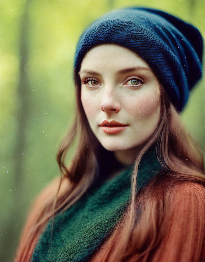 Freckled woman in blue hat and green scarf with subtle smile in blurred greenery background