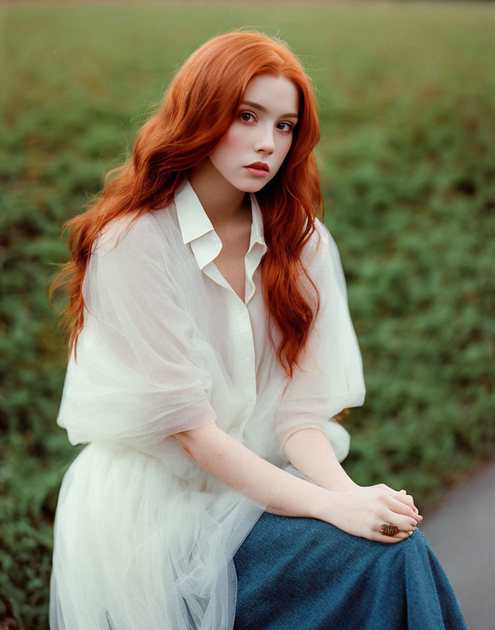 Vibrant red-haired woman in white blouse and blue skirt gazes at camera in green field