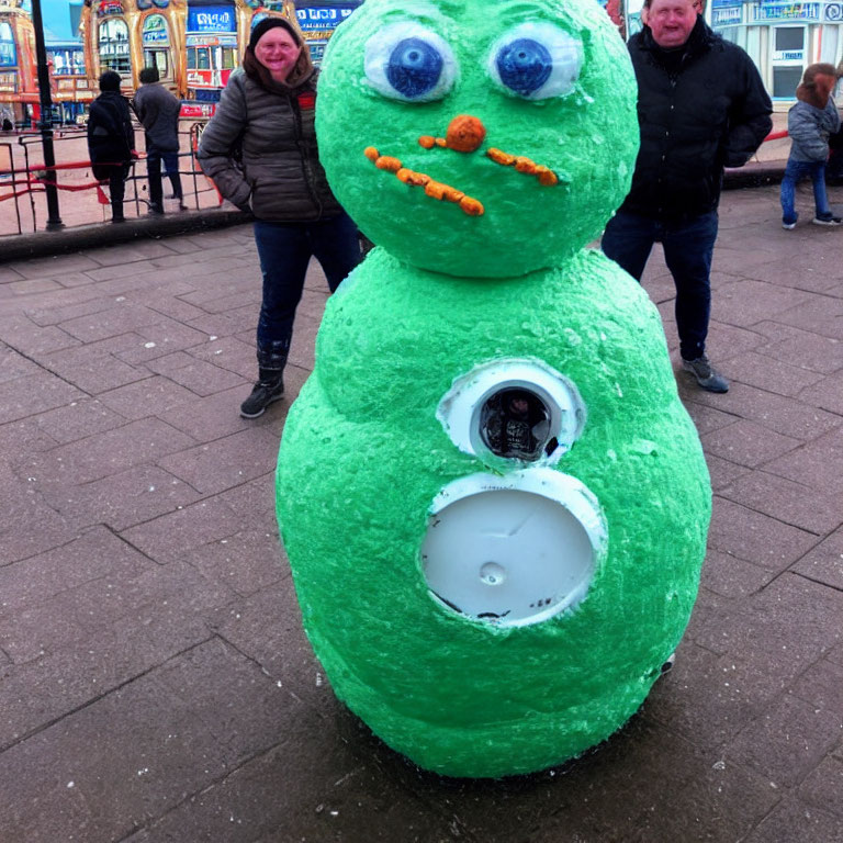 Green snowman with googly eyes, carrot nose, and white lid mouth, with two people behind
