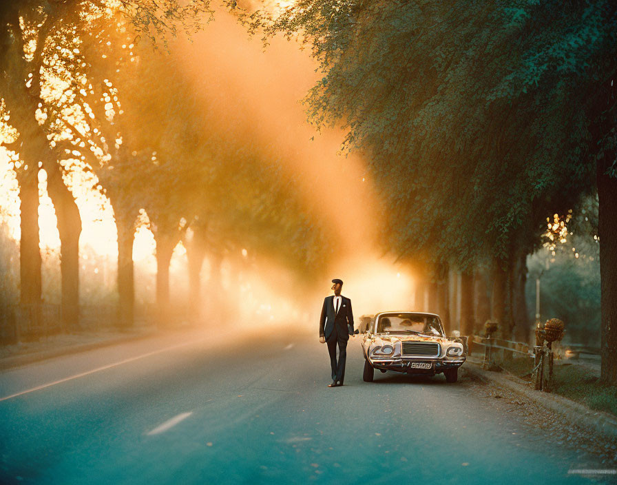 Man in suit on tree-lined road at sunset with classic car and headlights.