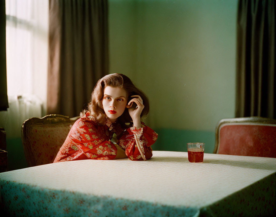 Pensive woman in red blouse at table near window with sheer curtains