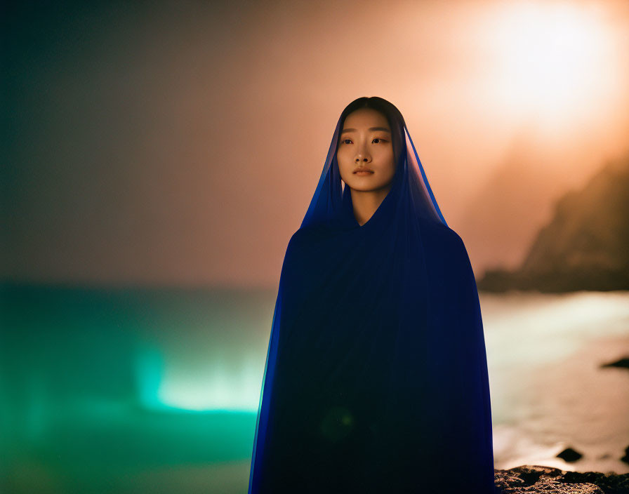 Mysterious figure in blue cloak by bioluminescent shore at sunset