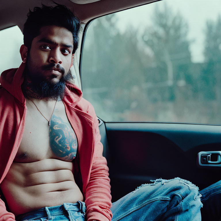 Tattooed man in red hoodie and ripped jeans sitting in car