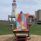 Colorful Rainbow Adirondack Chair on Pebble Beach with Lighthouse and Rainbow Sky