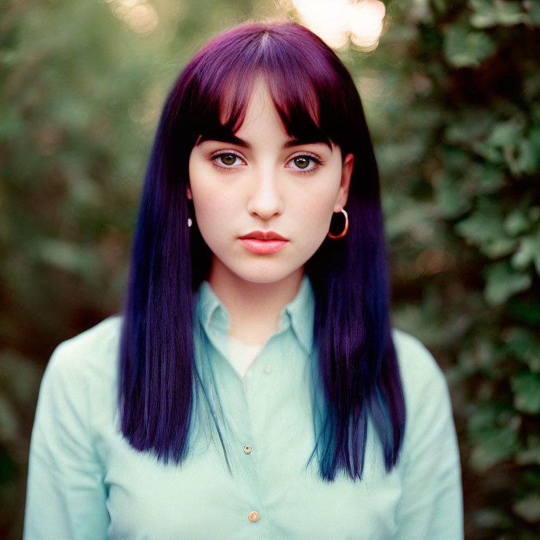 Purple-haired woman in mint-green blouse gazes at camera with blurred foliage background