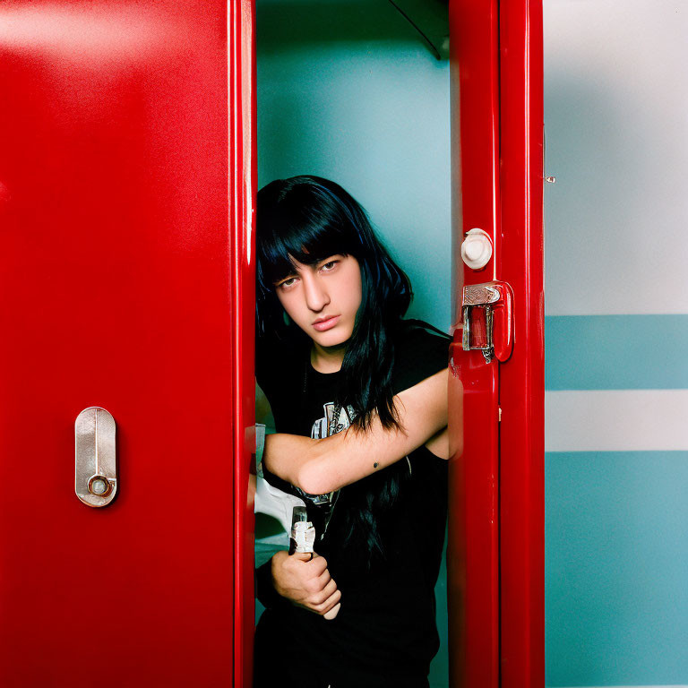 Serious person with black hair and fringe behind vibrant red door