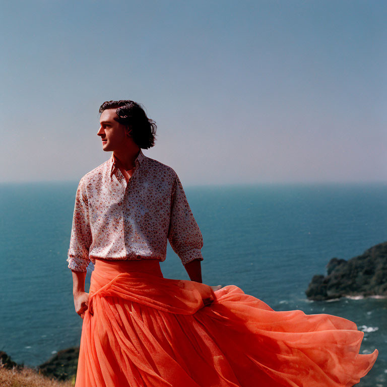 Person in patterned shirt and orange skirt gazes at sea from coastal overlook