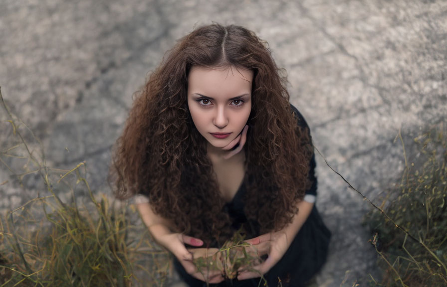 Curly-haired woman sitting on ground surrounded by grass on textured surface