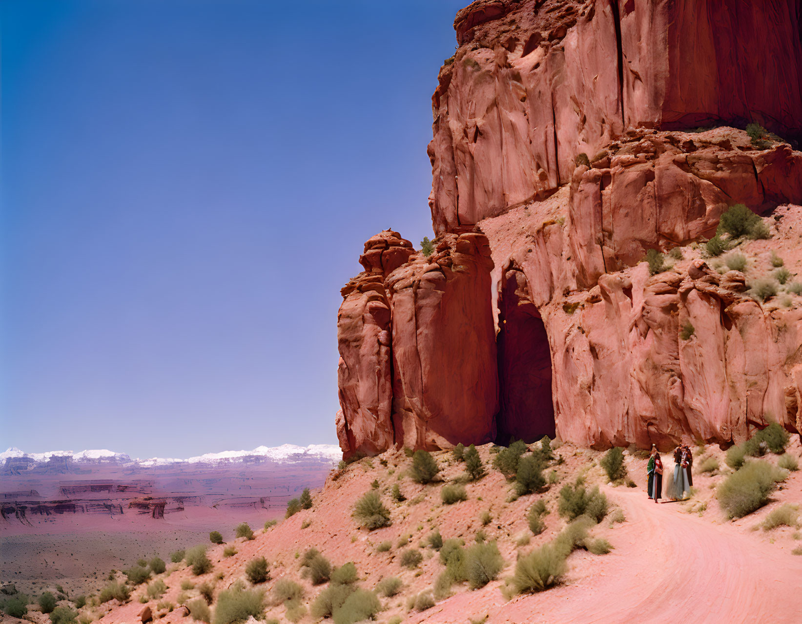 Red dirt path by towering red rock cliffs with distant snow-capped mountains and two people walking