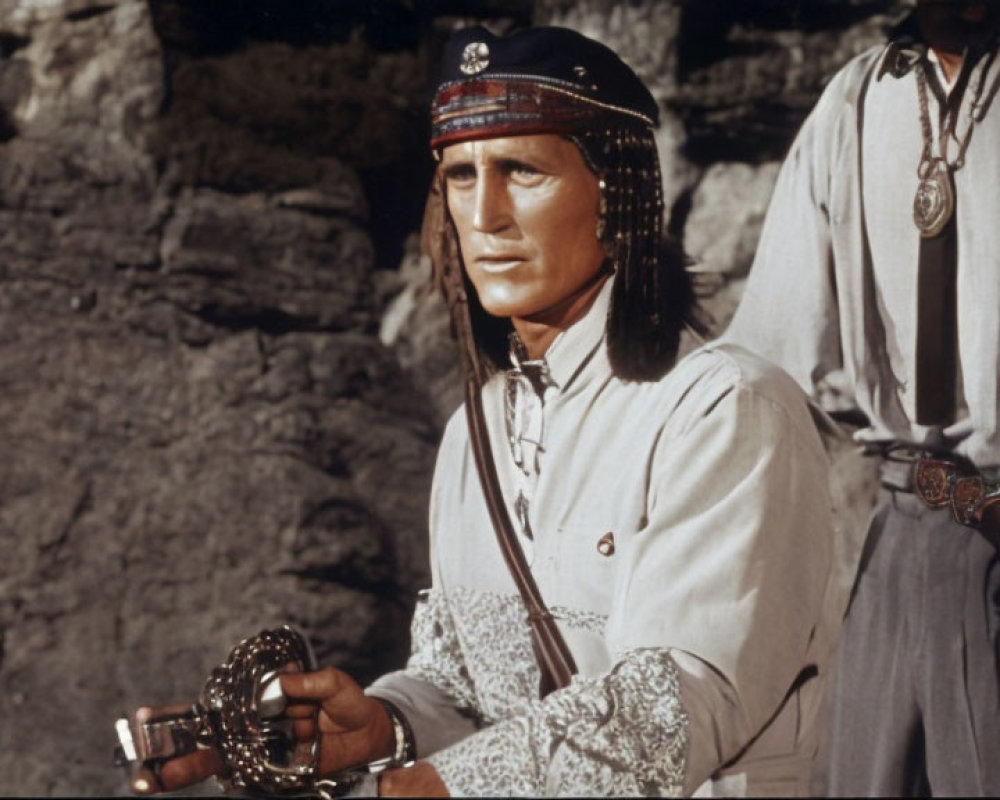 Person in traditional clothing holds shackles with rocky backdrop and another figure visible