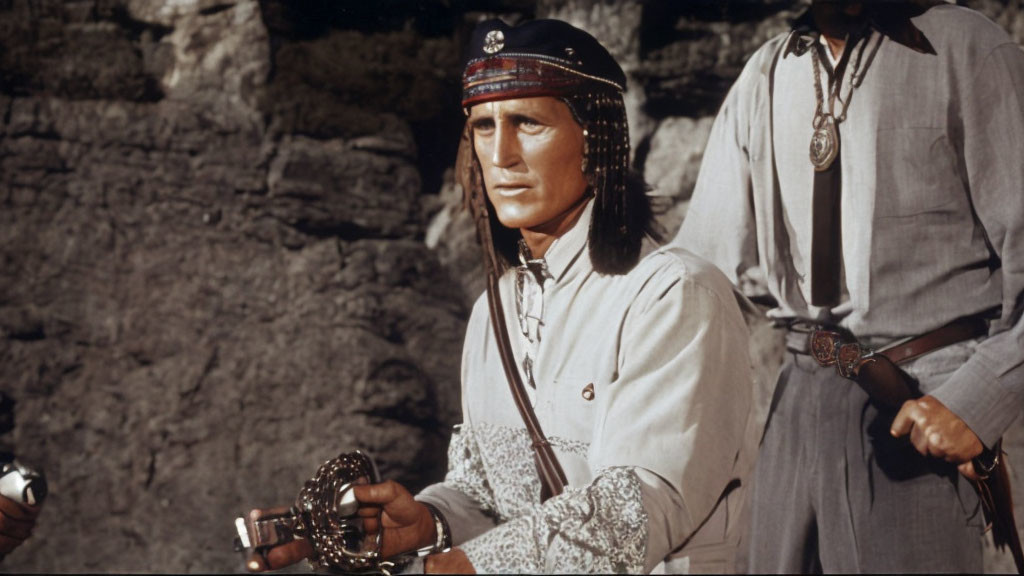 Person in traditional clothing holds shackles with rocky backdrop and another figure visible
