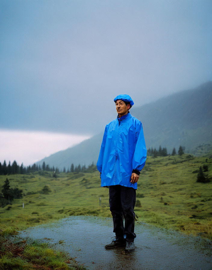 Man in Blue Raincoat Standing on Wet Path with Misty Green Hills