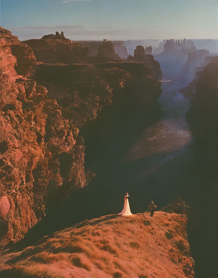 Bride in long dress on cliff overlooking canyon with river, photographer capturing moment