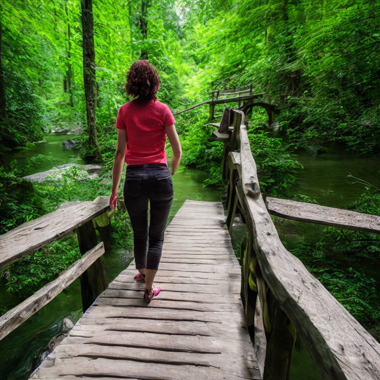 Woman in red top and dark pants crossing wooden bridge over stream in lush forest