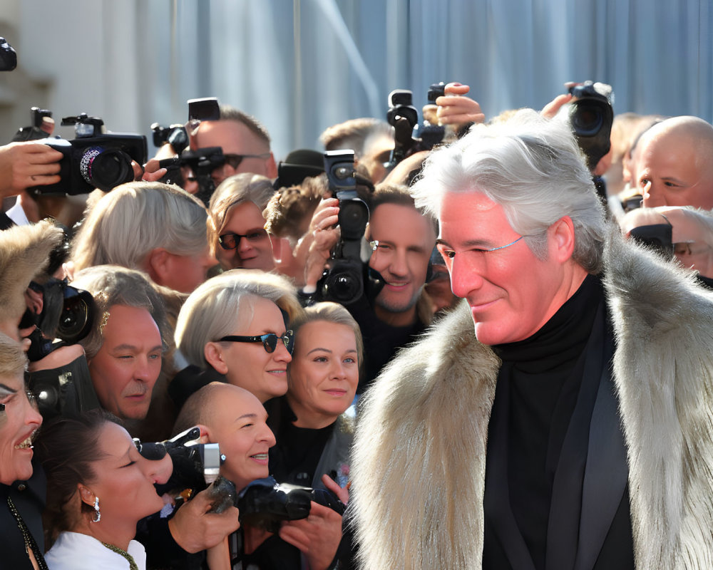 Silver-haired man in fur coat and turtleneck smiles amid photographers.