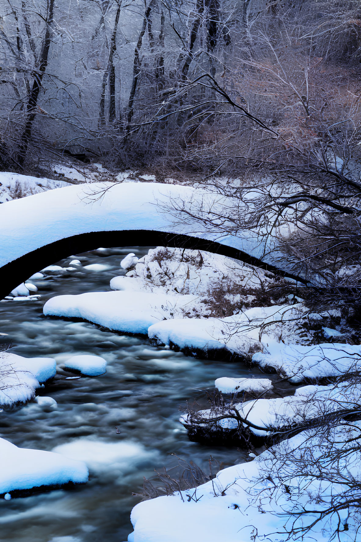 Snow-covered arched stone bridge over gently flowing river in serene winter scene