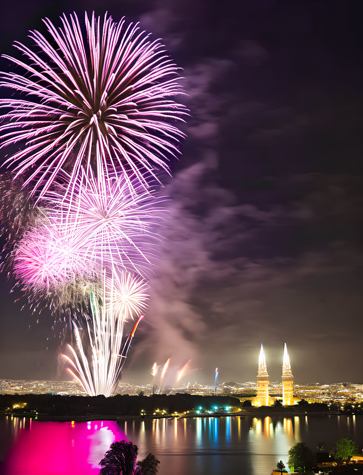 Colorful fireworks light up night cityscape with illuminated towers and reflections on water