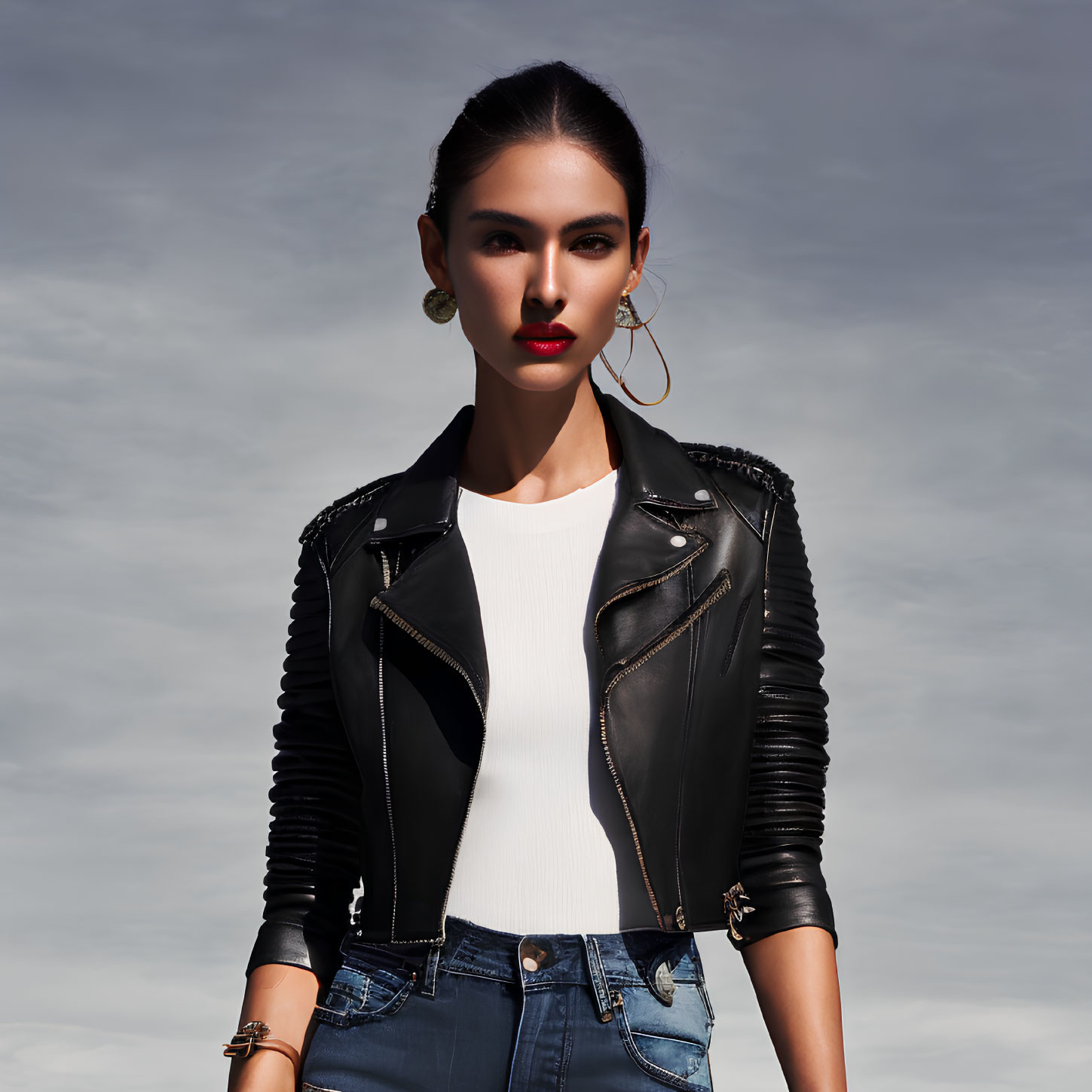 Confident woman in white top, leather jacket, jeans, and hoop earrings against cloudy sky