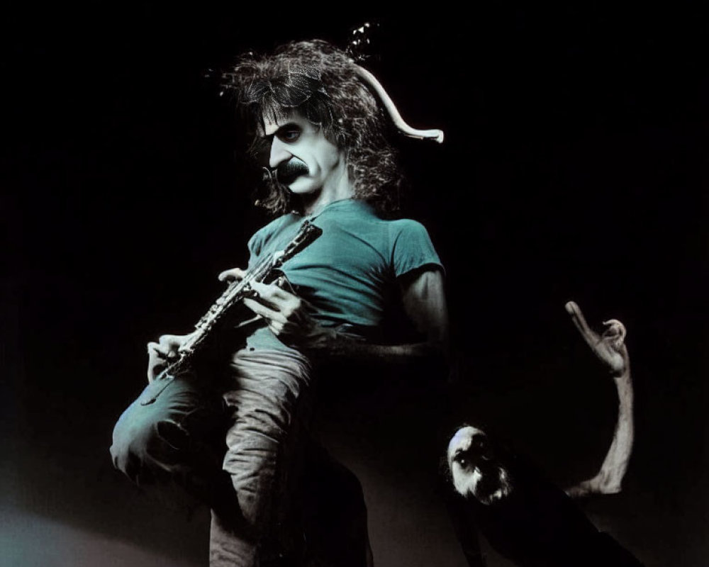 Mustached musician with curly hair plays guitar on stage with dramatic lighting, audience member's hand visible