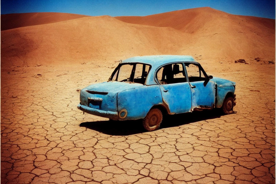 Abandoned blue car on cracked dry ground with sand dunes and clear sky