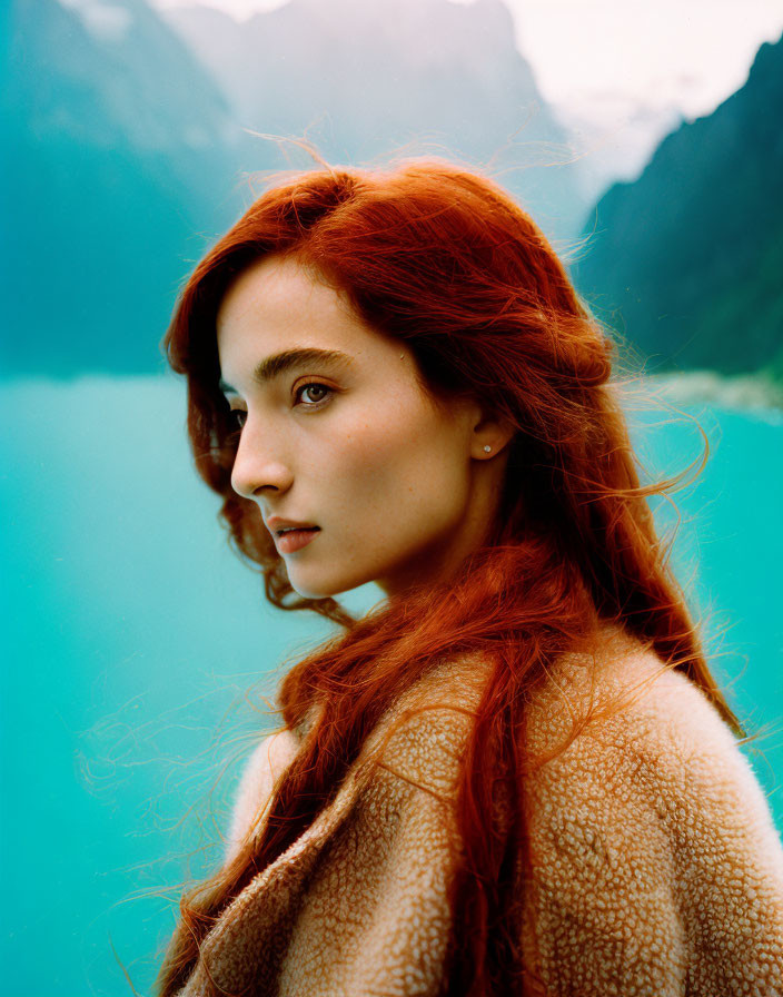 Red-haired woman with fair skin looking over shoulder by turquoise lake and green mountains
