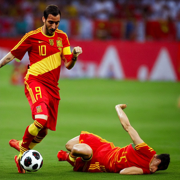 Soccer players in red and yellow kits on pitch: one standing, one fallen