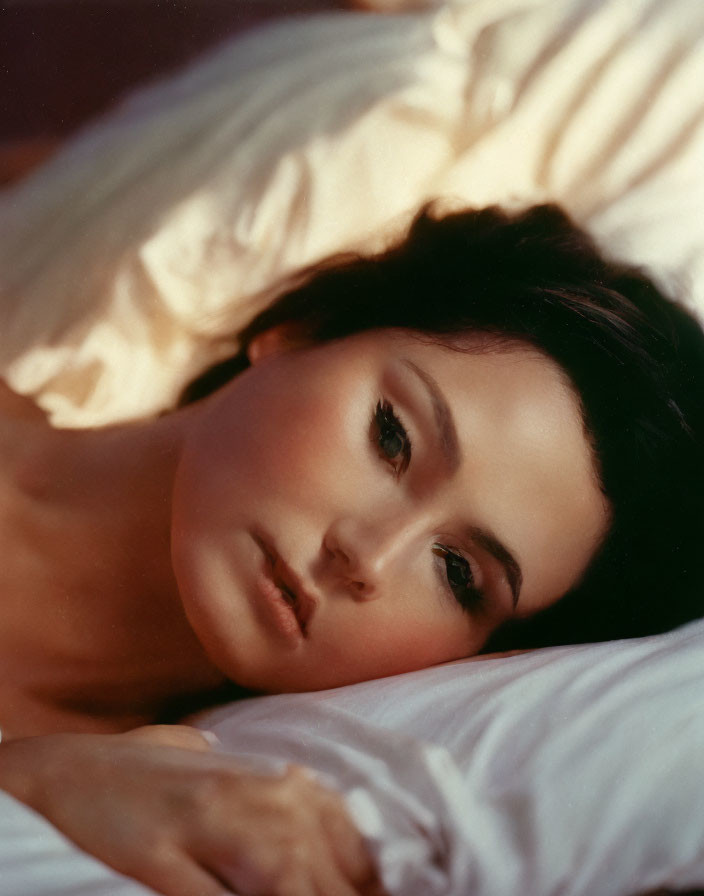 Dark-haired woman lying on white pillow gazes thoughtfully.