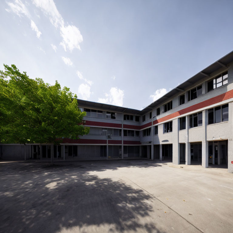 Two-story U-shaped building in empty school courtyard under blue sky with large green tree.